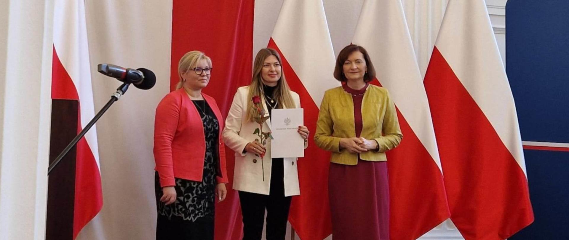 Na tle biało-czerwonych flag stoją od lewej: Małgorzata Dankowska - dyrektor Wydziału Polityki Społecznej Podkarpackiego Urzędu Wojewódzkiego, Katarzyna Palider - nagrodzona pracownica Gminnego Ośrodka Pomocy Społecznej w Jaśle z dyplomem w dłoni, Ewa Leniart - Wojewoda Podkarpacki