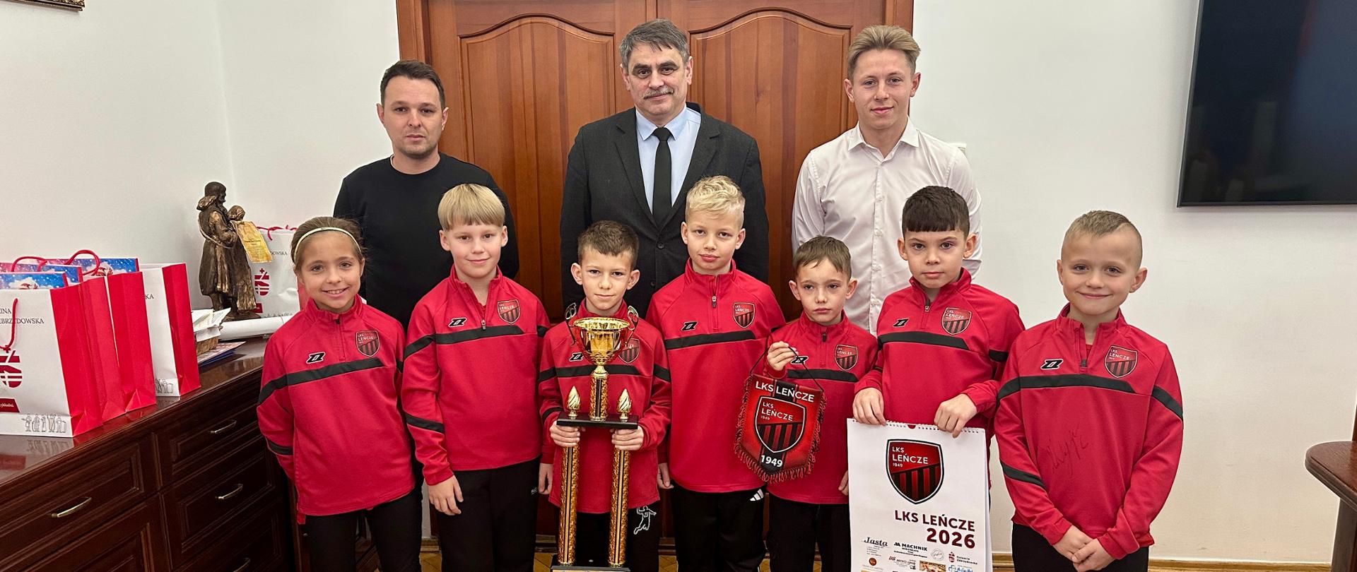 Grupowe zdjęcie młodych piłkarzy drużyny LKS Leńcze w czerwonych strojach klubowych, stojących w gabinecie urzędowym na parkietowej podłodze. Dzieci ustawione są w pierwszym rzędzie, jedno z nich trzyma duży złoty puchar, a inne prezentuje klubowy proporzec oraz kalendarz LKS Leńcze na 2026 rok. Za zawodnikami stoją trzej dorośli mężczyźni w strojach wizytowych i casualowych. W tle widoczne są drewniane drzwi, meble biurowe, telewizor oraz elementy wystroju urzędu. Zdjęcie dokumentuje uroczyste spotkanie po sportowym sukcesie drużyny.