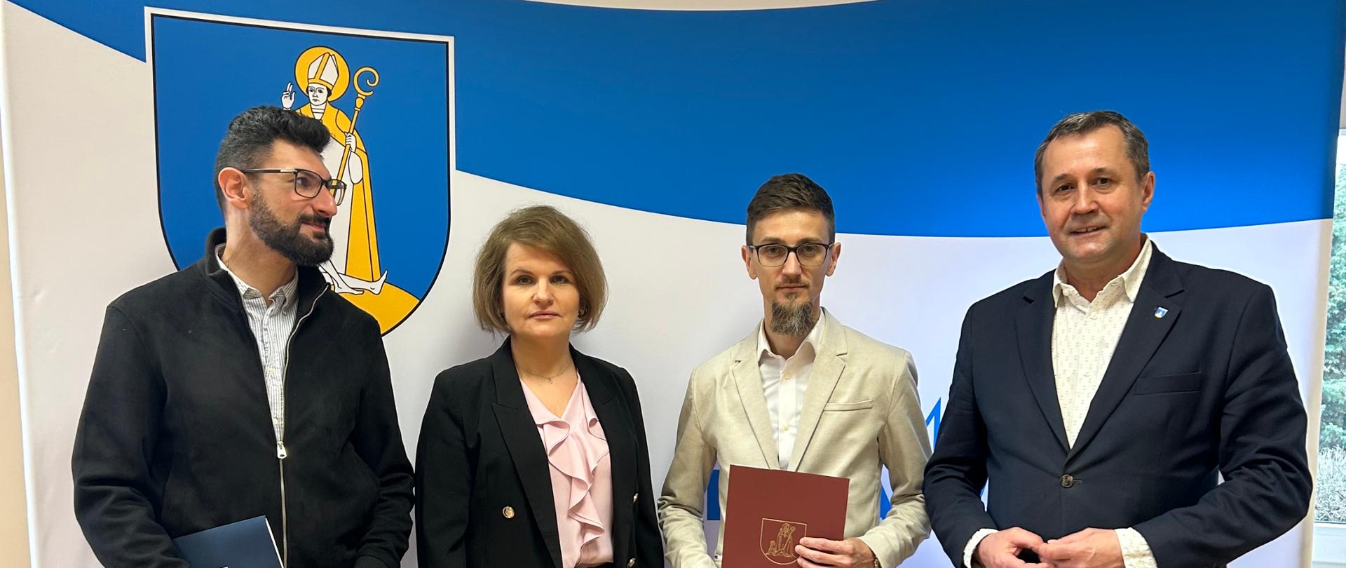 Cztery osoby stoją w pomieszczeniu przed ścianką z herbem i kolorami niebiesko-żółtymi. Dwie osoby trzymają teczki z dokumentami, a pozostali stoją obok nich. Wszyscy są ubrani w stylu biznesowym i pozują do zdjęcia.