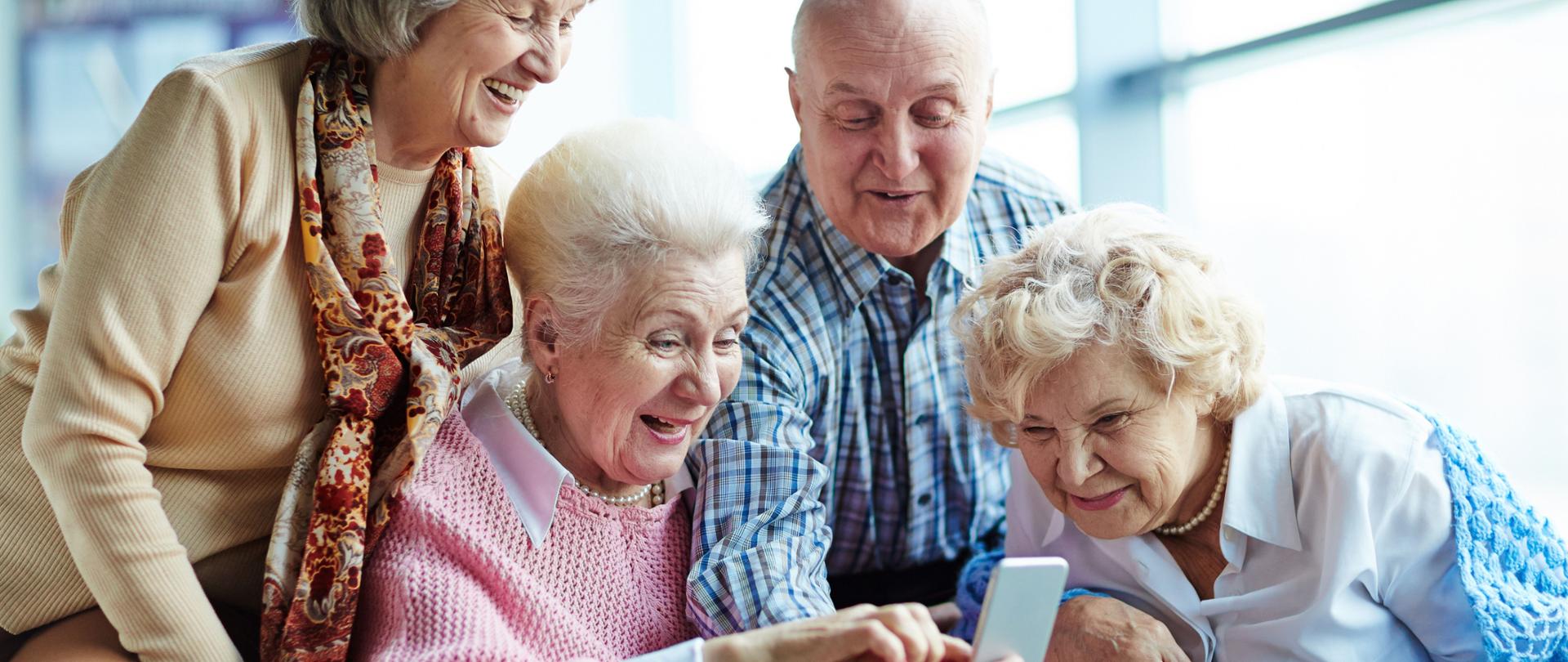 Na fotografii czwórka seniorów, dwie kobiety i jeden mężczyzna. Kobieta z prawej strony ubrana w białą koszulę oraz niebieską narzutkę. Obok niej mężczyzna ubrany w niebieską koszulę. Trzyma on w ręku telefon na którym pokazuje coś seniorkom. Obok niego starsza kobieta w różowym swetrze a koło niej kobieta w beżowym swetrze. Wszyscy są szczęśliwi i się uśmiechają. 