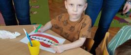 Dzieci w trakcie warsztatów - uczą się jak myć zęby z wykorzystaniem szczoteczek do zębów i grafik prezentującej szeroki uśmiech. 