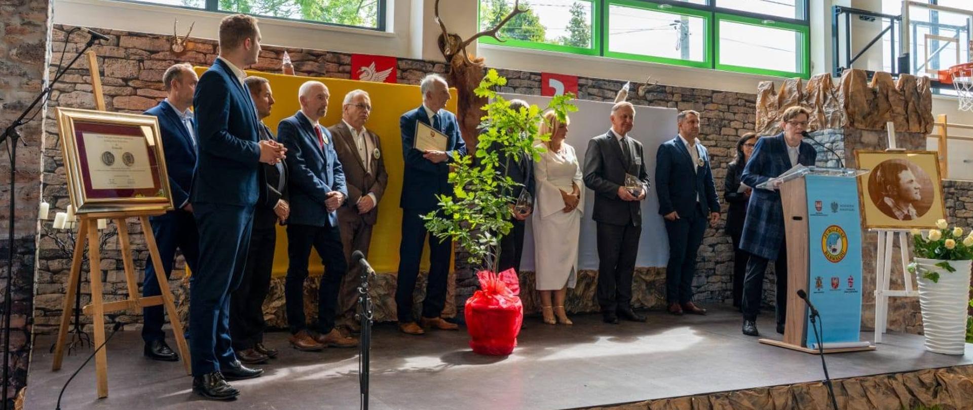 Na sali gimnastycznej szkoły w Barwałdzie. Na pierwszym planie zaproszeni goście. Burmstrz Kalwarii Zebrzydowskiej, Sotysi, Dyrektorzy oraz inni. Na fotografii widać medal jaki przyznano Sołectwu Barwad.
