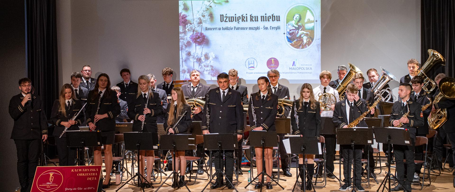 Na scenie stoi Kalwaryjska Orkiestra Dęta w pełnym składzie, ubrana w galowe, ciemne mundury. Muzycy trzymają instrumenty dęte – m.in. trąbki, klarnety, saksofony, flety i tuby. Przed nimi ustawione są pulpity nutowe. Po lewej stronie jeden z członków orkiestry przemawia do mikrofonu. W tle, na dużym ekranie, wyświetlony jest slajd z tytułem „Dźwięki ku niebu – koncert w hołdzie Patronce muzyki – św. Cecylii” oraz grafiką przedstawiającą św. Cecylię. Scena jest jasno oświetlona, a orkiestra prezentuje się przygotowana do koncertu.