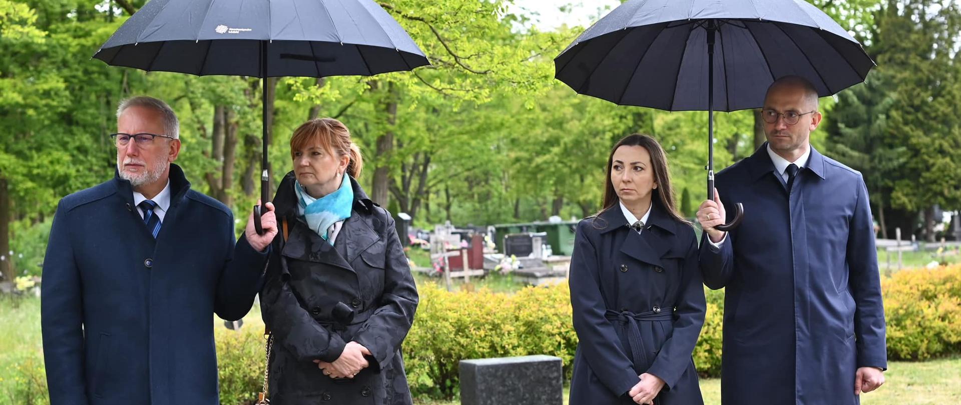 Władze miasta pod parasolami stoją na cmentarzu podczas uroczystości.