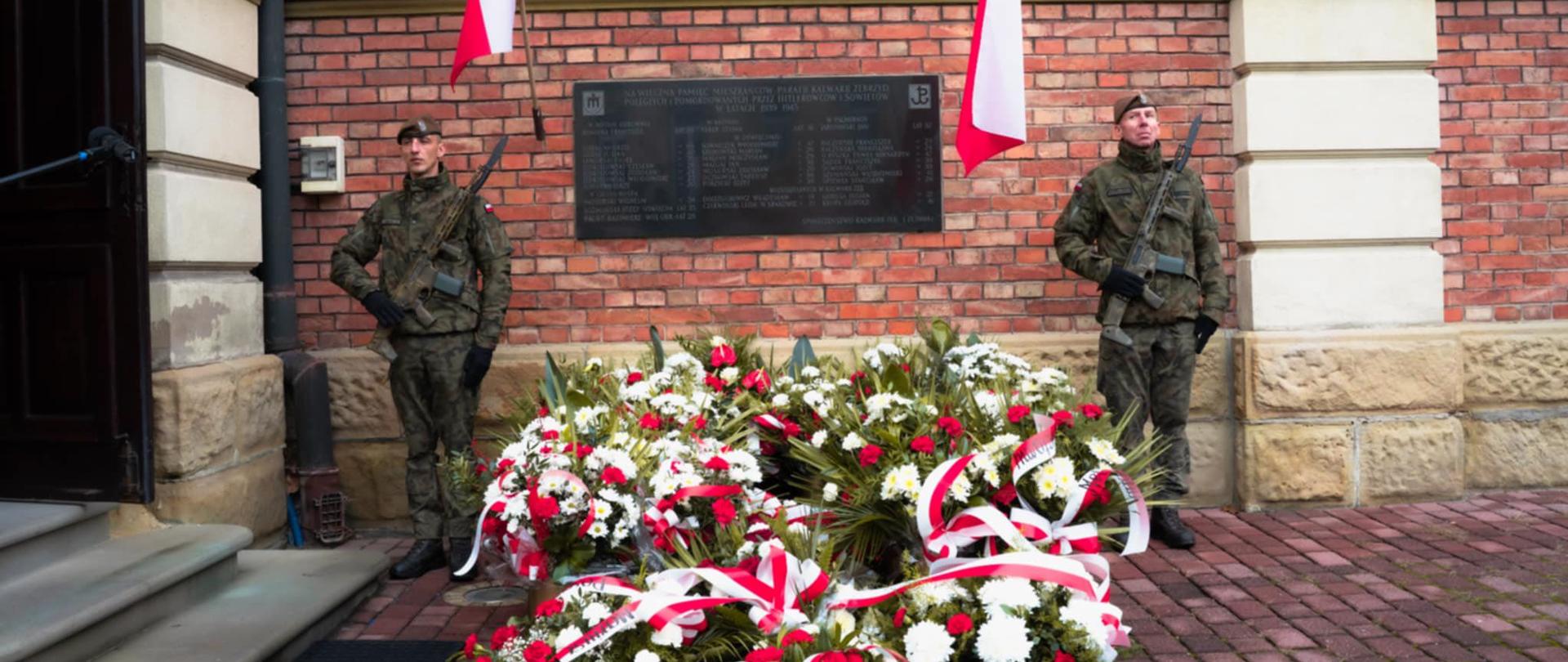 Dwóch żołnierzy w mundurach stoi na warcie przy tablicy pamiątkowej na ceglanym murze, przed wejściem do budynku. Przed nimi ułożone są liczne wieńce z biało-czerwonych kwiatów ozdobione wstęgami w barwach narodowych. Po obu stronach tablicy powiewają polskie flagi