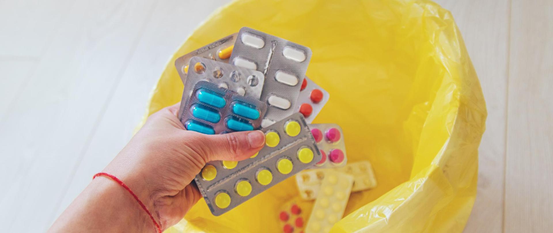 Dłoń trzyma kilka blistrów z tabletkami i kapsułkami nad otwartym żółtym workiem na odpady medyczne, do którego wyrzucane są przeterminowane lub niepotrzebne leki.