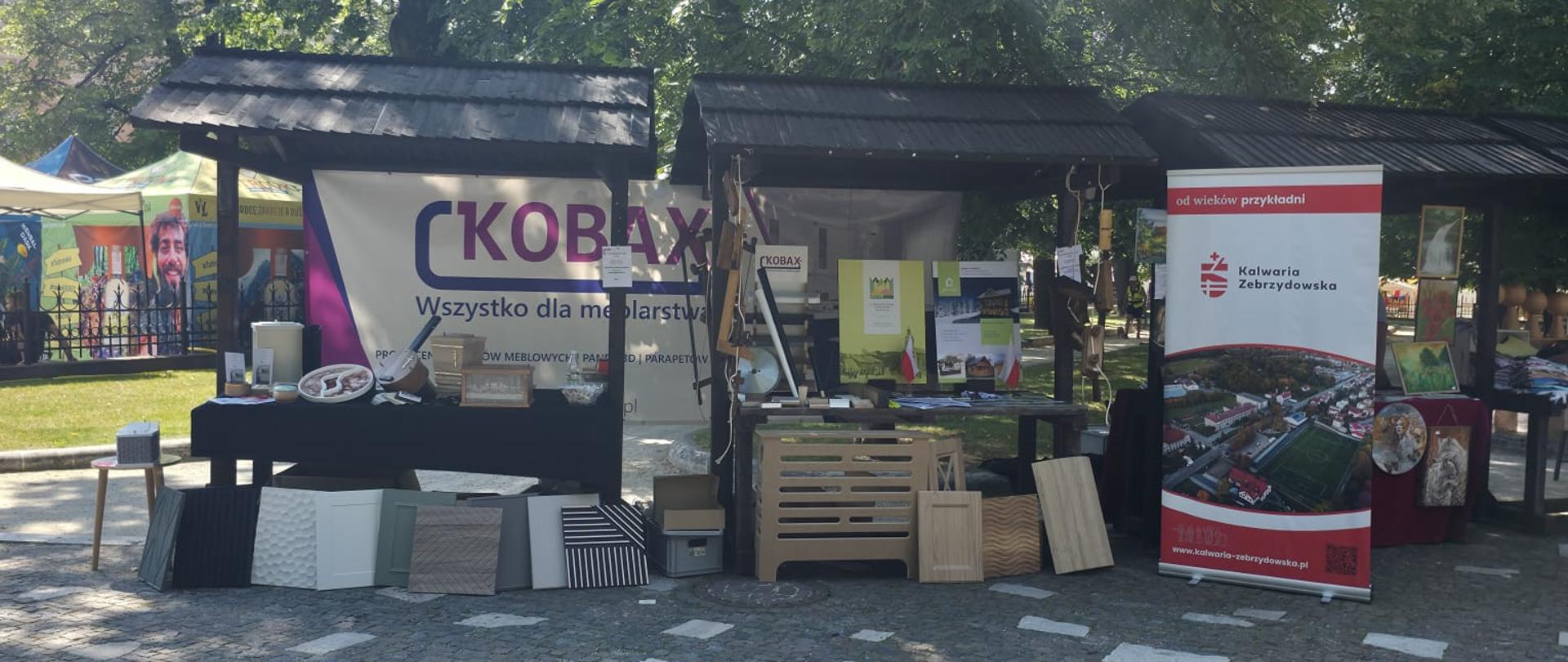 Stoisko wystawowe ustawione na brukowanym placu w cieniu drzew. Po lewej stronie widoczny jest baner z napisem KOBAX – Wszystko dla meblarstwa, prezentujący różne materiały i elementy wykończeniowe. Obok znajduje się stoisko z ekspozycją drewnianych wyrobów i materiałów promocyjnych, w tym roll-up Gminy Kalwaria Zebrzydowska z panoramą miasta i logiem. W tle widać kolejne kolorowe namioty oraz ludzi odwiedzających wydarzenie.