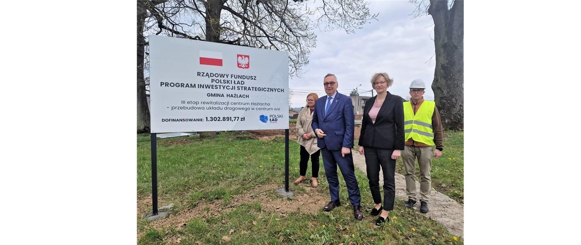 Wiceminister Stanisław Szwed w towarzystwie przedstawicieli władz Gminy Hażlach oraz głównego wykonawcy inwestycji. Cztery osoby stoją obok tablicy informującej o inwestycji.