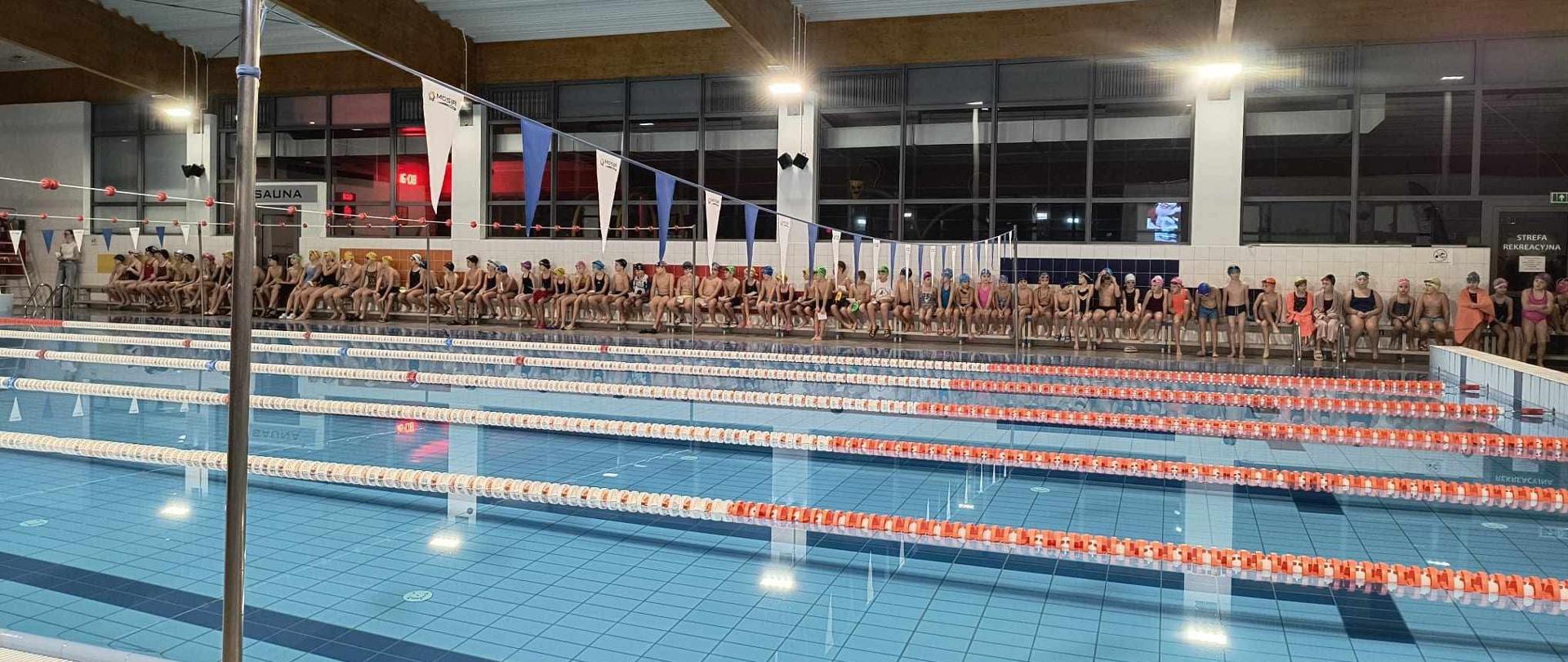 Na pierwszym planie widać nieckę z wodą, a dalej za nią siedzących na ławkach zawodników.