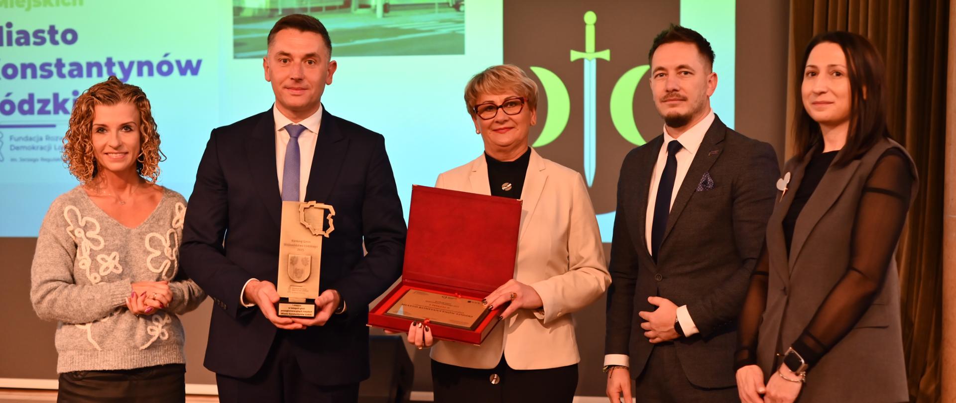 Burmistrz Konstantynowa Łódzkiego Robert Jakubowski oraz przewodnicząca rady miejskiej Jadwiga Czekajewska pozują z otrzymaną statuetką i tablicą podczas gali wręczenia nagród Rankingu Gmin Województwa Łódzkiego 2025; w tle prezentacja z logo miasta.