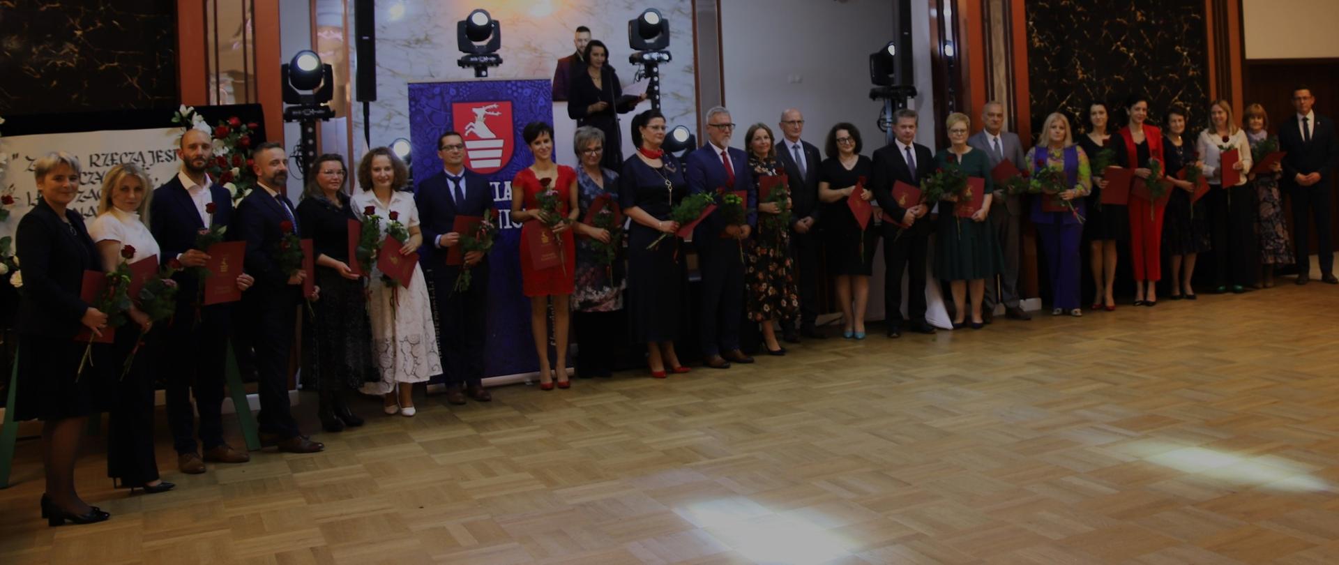 Nagrodzeni nauczyciele stojący w sali pod sceną i trzymający w rękach czerwone róże oraz listy gratulacyjne