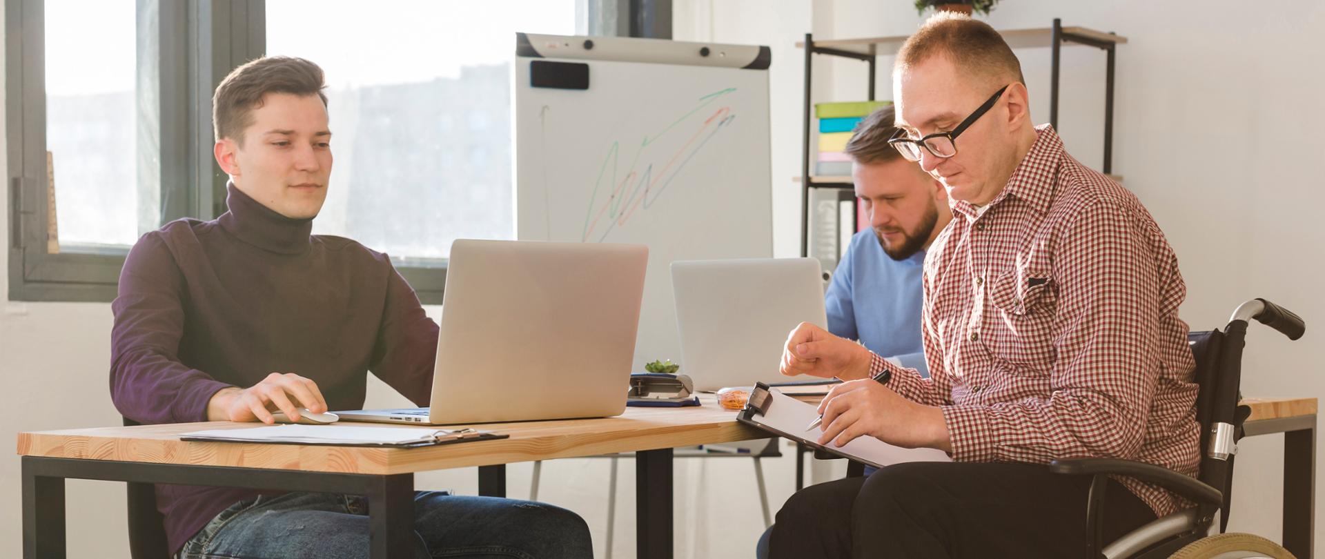 Trzech mężczyzn pracujących razem w biurze przy biurkach z laptopami. Jeden z nich siedzi na wózku inwalidzkim i przegląda dokumenty.