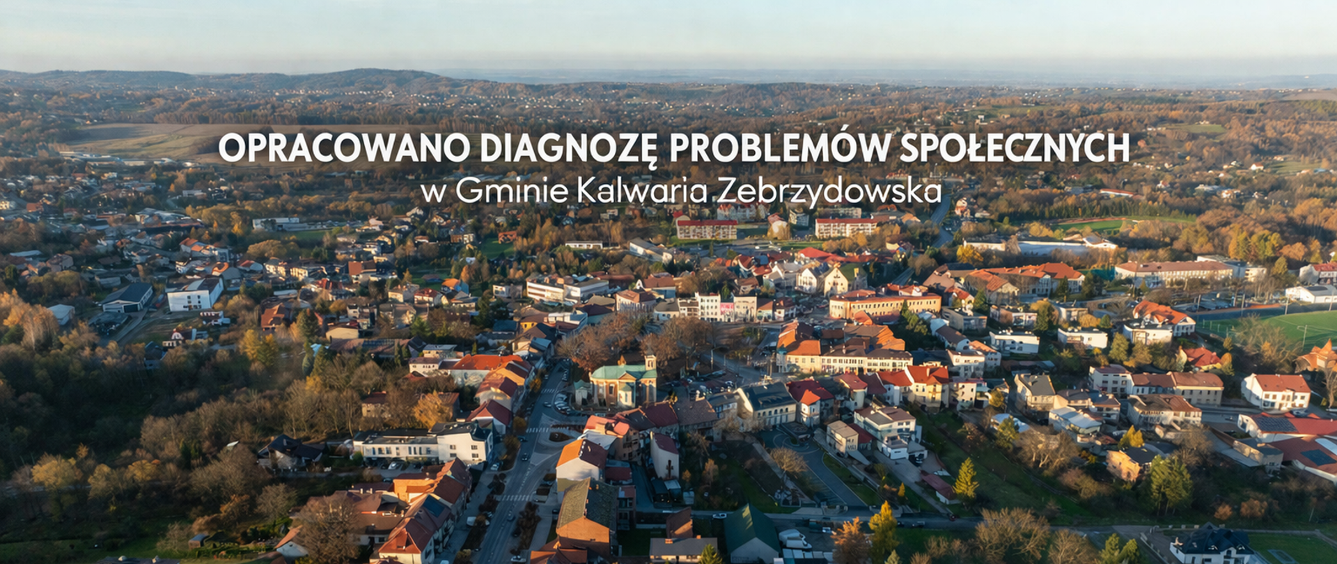 Panoramiczne ujęcie lotnicze Kalwarii Zebrzydowskiej z widoczną zabudową miejską, kościołem oraz otaczającymi wzgórzami. Na tle zdjęcia umieszczono napis: „Opracowano diagnozę problemów społecznych w Gminie Kalwaria Zebrzydowska”.