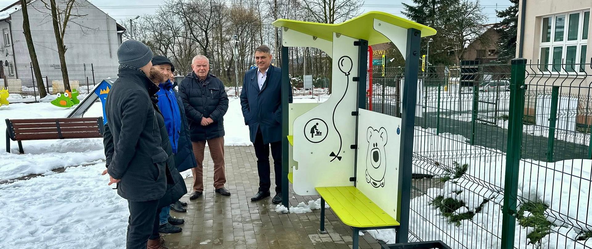 Grupa kilku mężczyzn stoi na terenie ośnieżonego placu zabaw przy szkole. Na pierwszym planie widać nowo zamontowaną ławkę z przewijakiem w jasnych kolorach, ozdobioną prostymi ilustracjami balonika i misia. W tle znajdują się ogrodzenie, budynek szkolny oraz elementy placu zabaw, a cała scena odbywa się w zimowej scenerii.