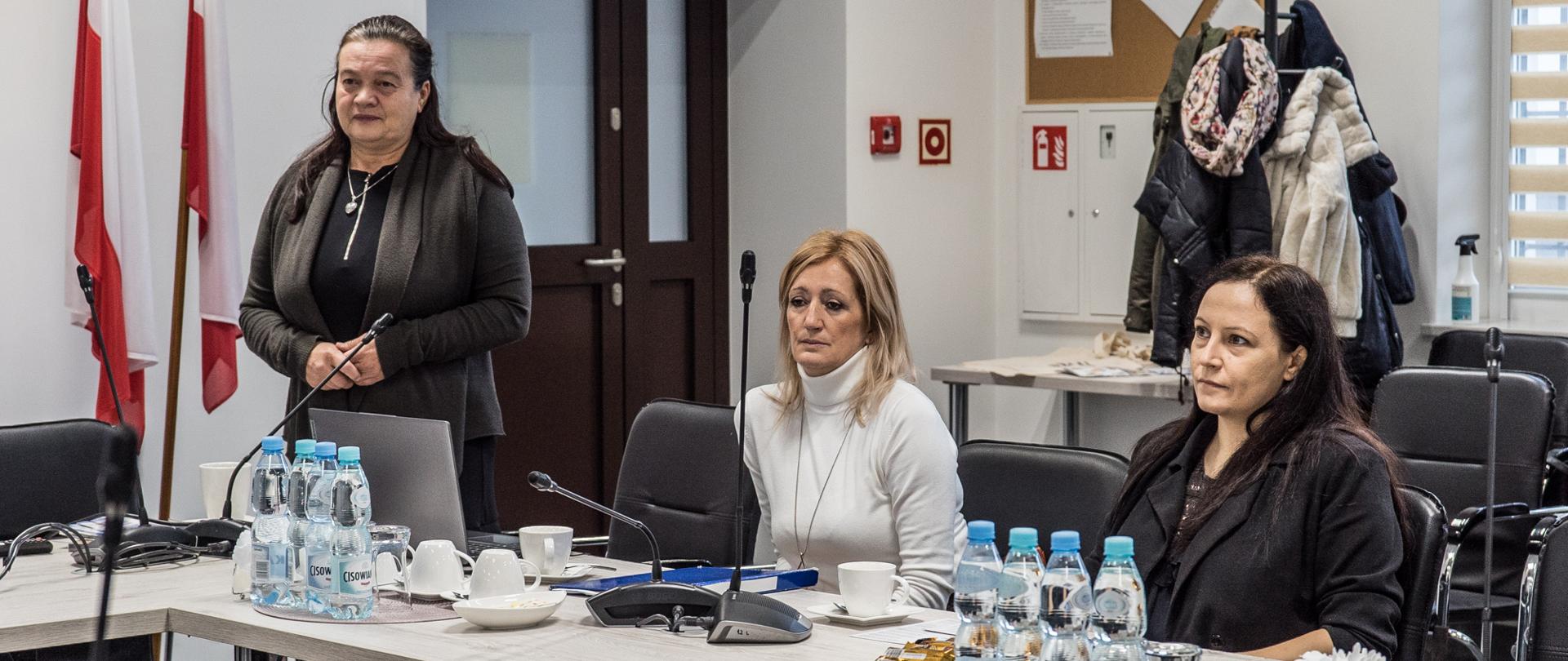 z lewej strony stoi kobieta w ciemnym stroju z założonymi rekami po prawej stronie siedzi kobieta w długich jasnych włosach i w jasnym swetrze, najbardziej z prawej strony siedzi kobieta o ciemnych włosach po ramiona w ciemnej marynarce 