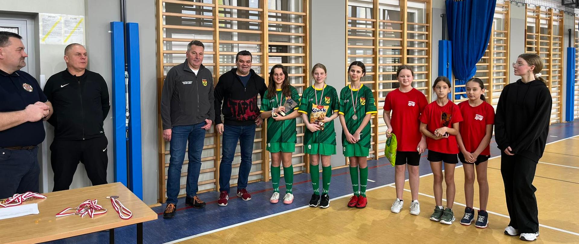 Na fotografii dziewczynki ubrane w stroje sportowe (trzy w stroje zielone oraz trzy w stroje pomarańczowo-czarne) oraz mężczyźni ubrani w ciemne stroje. Znajdują się na hali sportowej. Parkiet na którym stoją brązowo niebieski. Za osobami jasno brązowe drabinki. Z lewej Strony fotografii stolik, na którym leżą medale oraz nagrody.