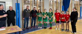 Na fotografii dziewczynki ubrane w stroje sportowe (trzy w stroje zielone oraz trzy w stroje pomarańczowo-czarne) oraz mężczyźni ubrani w ciemne stroje. Znajdują się na hali sportowej. Parkiet na którym stoją brązowo niebieski. Za osobami jasno brązowe drabinki. Z lewej Strony fotografii stolik, na którym leżą medale oraz nagrody.