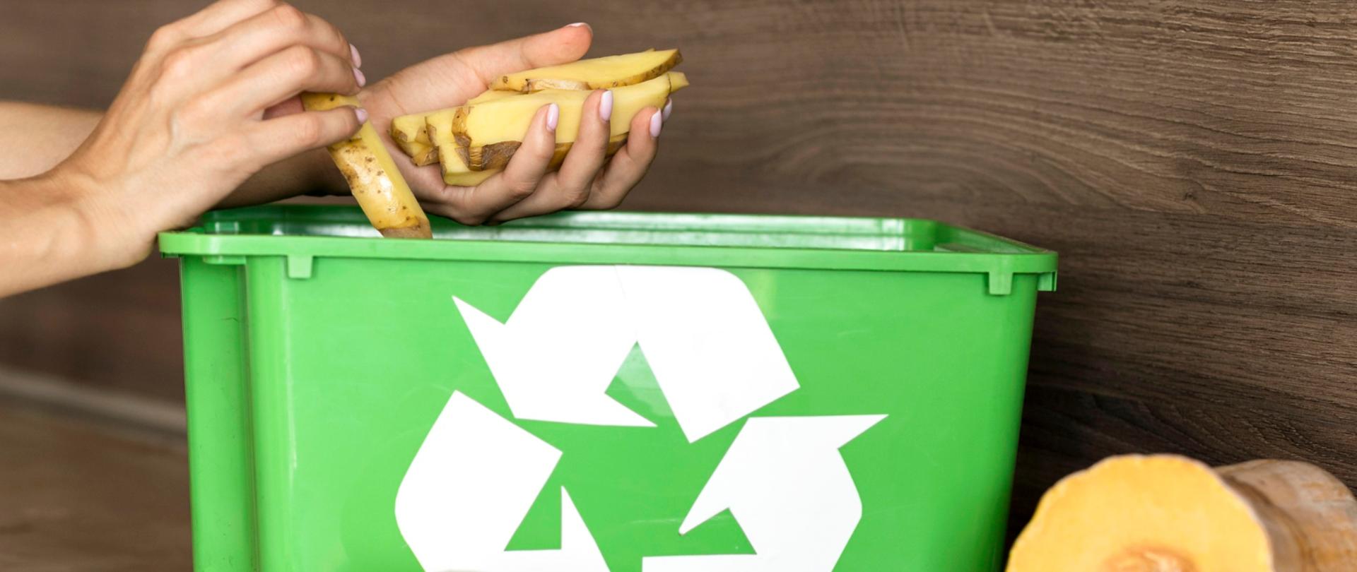 Zbliżenie na zielony pojemnik na bioodpady z symbolem recyklingu. Nad pojemnikiem dłonie wrzucają obierki z ziemniaków. Na blacie obok znajdują się produkty spożywcze i resztki organiczne, m.in. jajka, ogórki, pomidor, cytrusy i przekrojona dynia, ilustrujące prawidłową segregację bioodpadów.