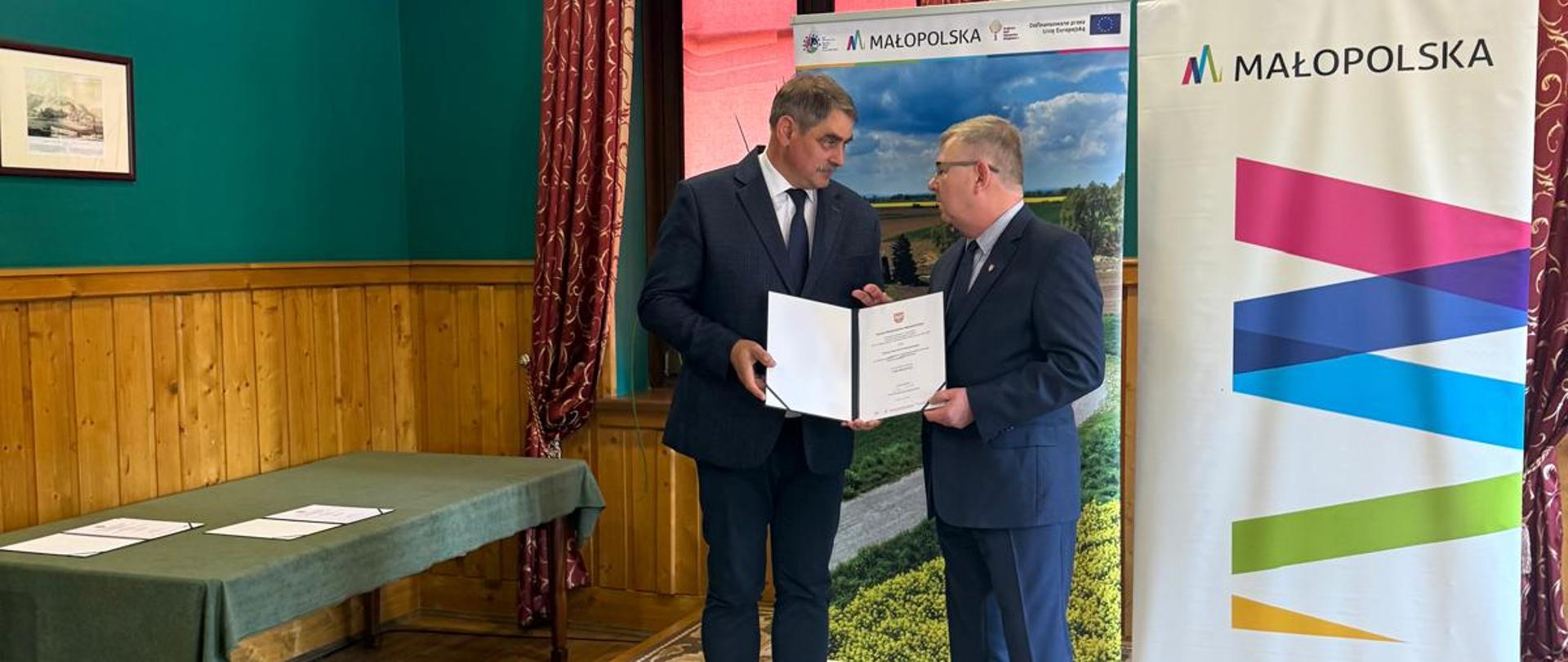 Dwóch mężczyzn w garniturach prezentuje podpisany dokument podczas oficjalnego spotkania. W tle znajdują się roll-upy z logotypem Małopolski oraz informacją o dofinansowaniu ze środków europejskich.