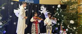 Kadr z występu 3 dzieci na scenie dwoje mniejszych gra na skrzypcach w strojach góralskich, starszy przebrany za anioła chłopak śpiewa kolędę. Scenografia zimowa, choinki, śnieg. 
