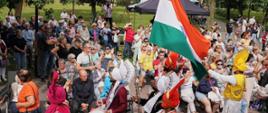 Artyści z Indii w kolorowych tradycyjnych strojach i turbanach wykonują taniec ludowy z użyciem instrumentów przed liczną publicznością w parku. Jeden z tancerzy niesie dużą flagę Indii.