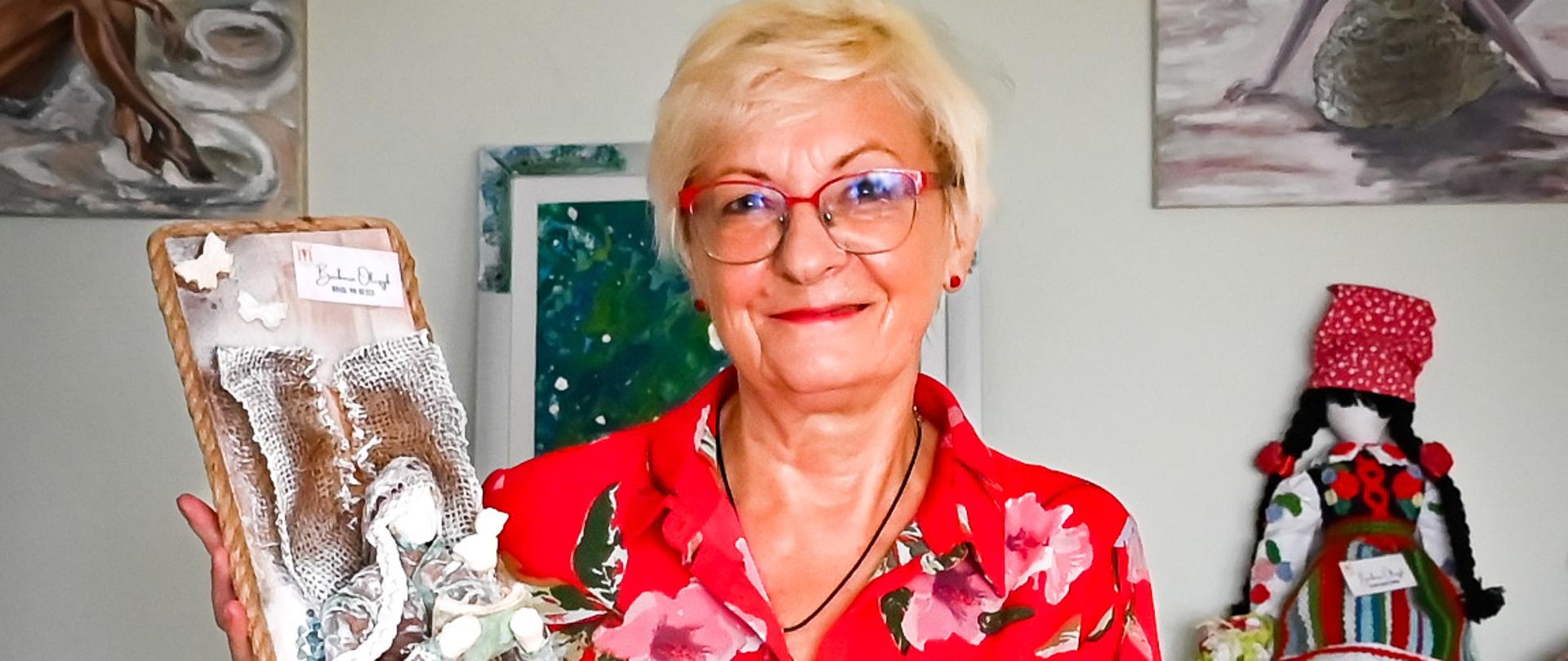 Kobieta w czerwonej sukience w kwiaty prezentuje własnoręcznie wykonaną dekorację artystyczną; w tle obrazy i rękodzieło.