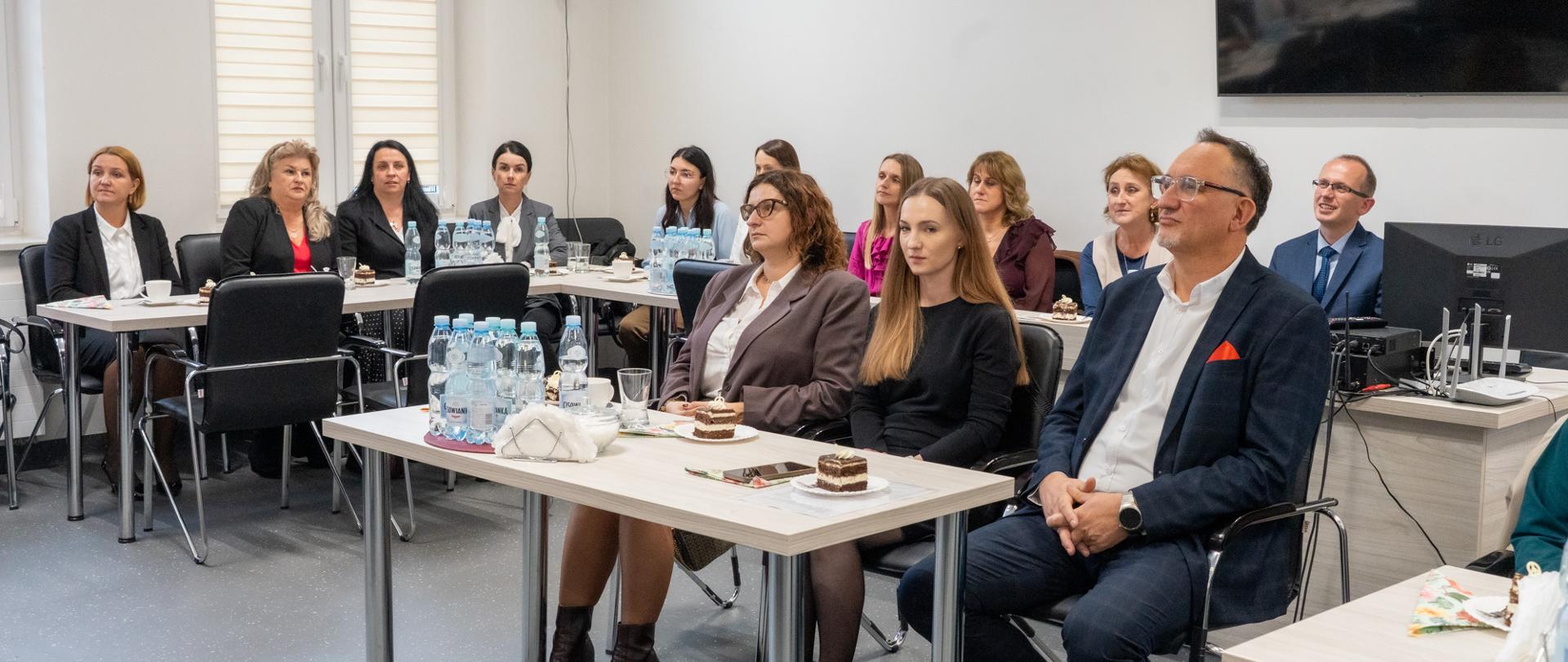 Zdjęcie przedstawia uczestników uroczystości wręczenia Nagród Burmistrza Miasta Kalwarii Zebrzydowskiej dla dyrektorów i nauczycieli, zorganizowanej z okazji Dnia Edukacji Narodowej. Uczestnicy siedzą przy stołach w sali konferencyjnej, przysłuchując się wystąpieniu. Na stołach znajdują się napoje, filiżanki oraz poczęstunek.