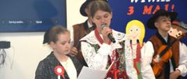 Występ muzyczny dwóch dziewczyn. Wyższa ubrana w strój góralski trzyma mikrofon i śpiewa, niższa trzyma tekst piosenki w ręku ubrana w strój galowy. W tle widać zespół góralski oraz dekoracje wiwat 3 maja.