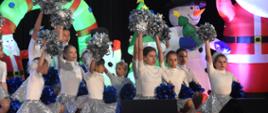 Grupa dziewcząt podczas występu na scenie. Mają srebrno-białe kostiumy. W rękach trzymają pompony.
