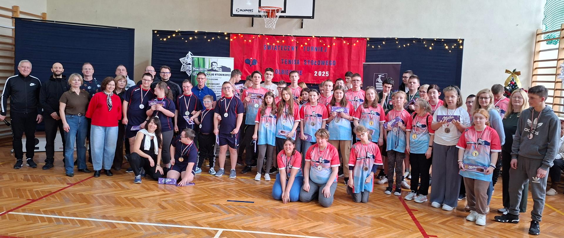 Uczestnicy turnieju z medalami na szyjach i czekoladami w rękach stoją na sali gimnastycznej wraz z opiekunami, dyrekcją Specjalnego Ośrodka Szkolno - Wychowawczego w Olbięcinie i Wiceprzewodniczącym Rady Powiatu w Kraśniku Michałem Stawiarskim.
