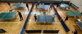 Widok z góry na halę gimnastyczną. Zawodnicy rozgrywają pojedynki w hali sportowej przy kilku rozstawionych stołach pingpongowych.