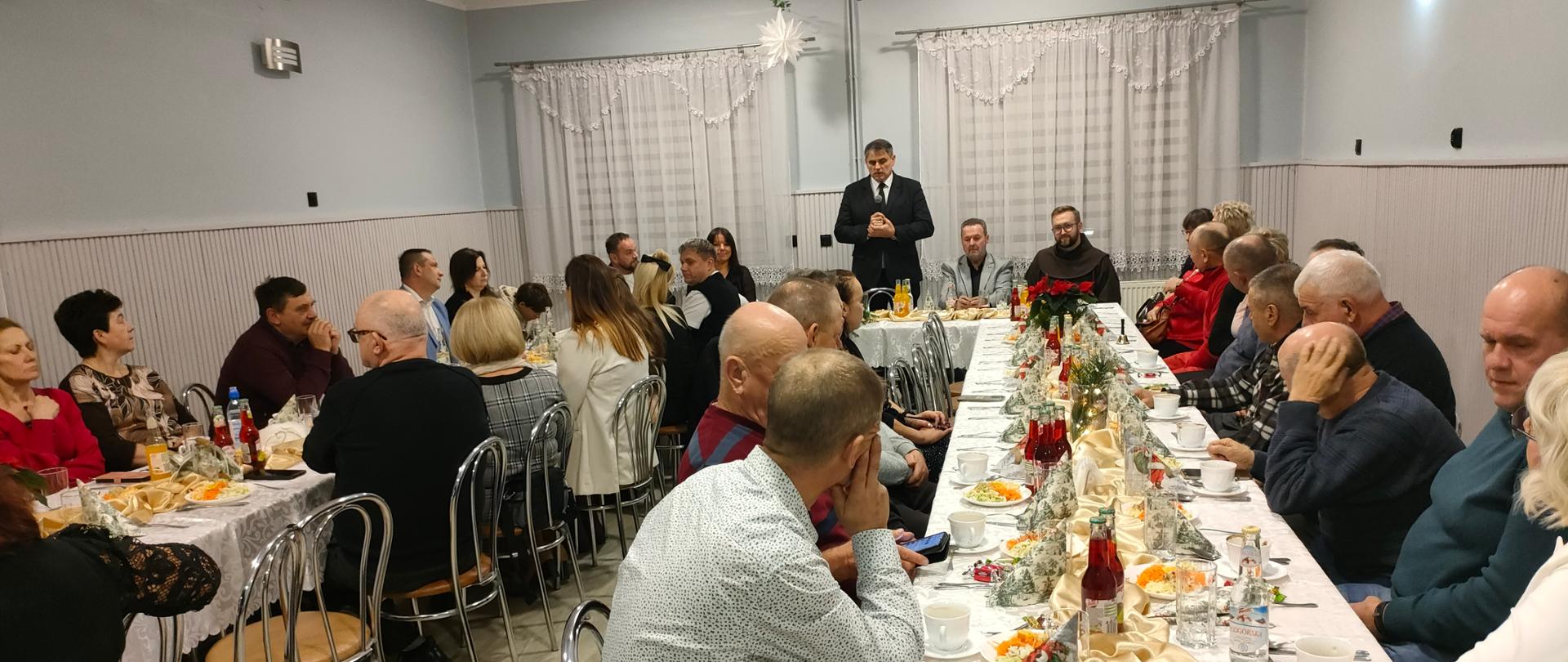 Spotkanie wigilijne przy długich, wspólnie zestawionych stołach w sali świetlicowej. Kilkadziesiąt osób siedzi przy białych obrusach, na których znajdują się tradycyjne potrawy, napoje i świąteczne dekoracje. W centralnej części sali stoi mężczyzna w garniturze, przemawiający do uczestników spotkania. Wnętrze jest jasno oświetlone, udekorowane subtelnymi ozdobami świątecznymi, a atmosfera wydarzenia ma uroczysty i wspólnotowy charakter.