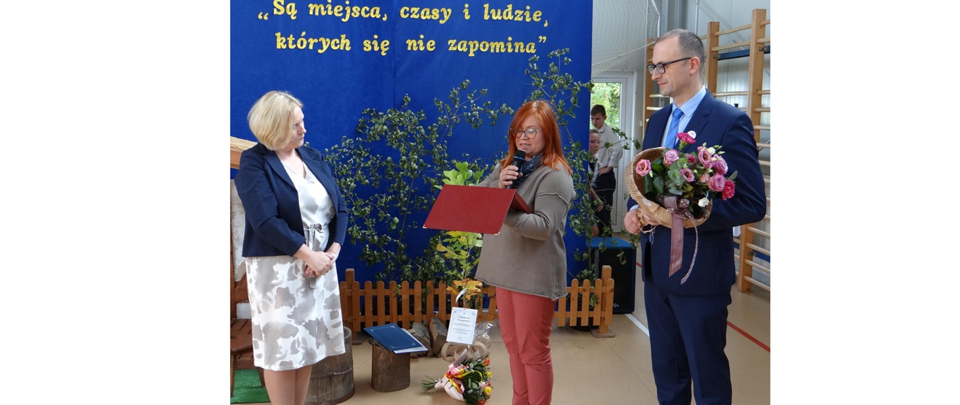 Dyrektor szkoły Aneta Orzyszek odbiera jubileuszowe życzenia od przedstawicieli władz Gminy Hażlach. Na pierwszym planie trzy osoby, w tym kobieta odczytująca życzenia z dyplomu oraz mężczyzna trzymający wiązankę kwiatów