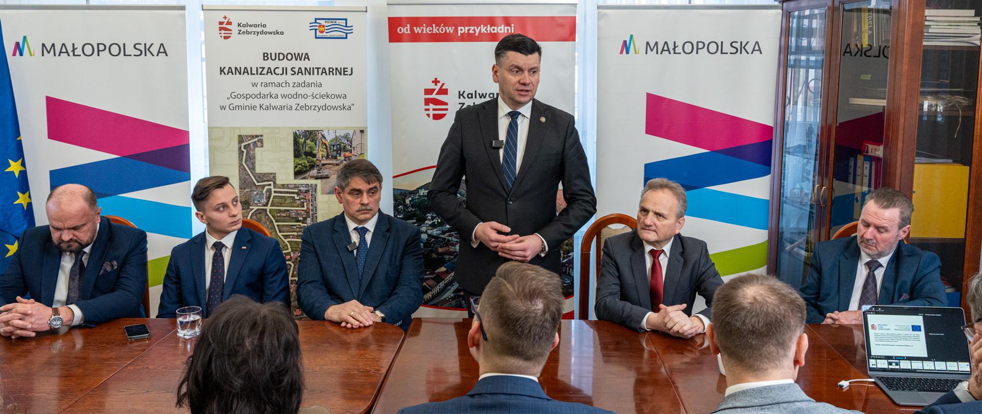 W sali konferencyjnej Urzędu Miasta Kalwarii Zebrzydowskiej trwa spotkanie dotyczące realizacji projektu budowy kanalizacji sanitarnej. Przy drewnianym stole siedzą samorządowcy i zaproszeni goście, a w centralnym punkcie stoi mężczyzna w garniturze przemawiający do zgromadzonych. W tle widoczne są roll-upy z logotypami Małopolski i Gminy Kalwaria Zebrzydowska oraz plansza informująca o inwestycji w gospodarkę wodno-ściekową. Po prawej stronie znajduje się przeszklona szafa z dokumentami, a za stołem siedzą uczestnicy spotkania, słuchający wystąpienia.