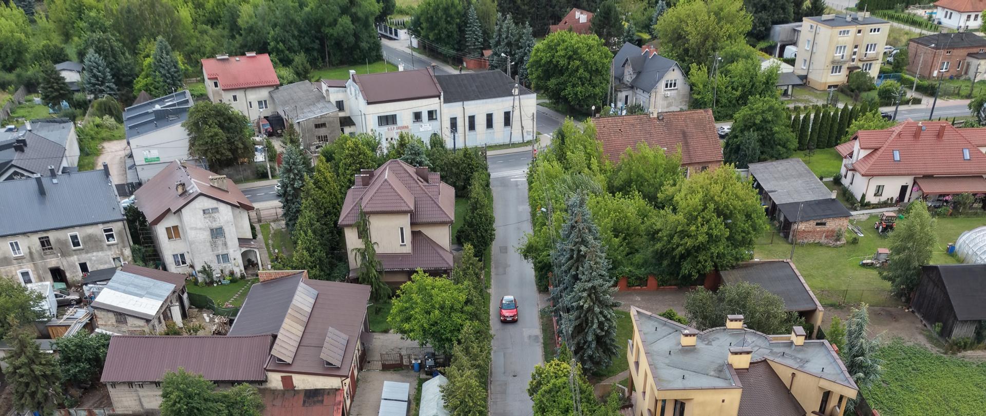 Widok z lotu ptaka na ulicę Warzywniczą przed przebudową. Wąska jezdnia przebiega przez osiedle domów jednorodzinnych. Po obu stronach ulicy widoczne są posesje z ogrodami, drzewa, samochody oraz zabudowania gospodarcze i tunele foliowe.