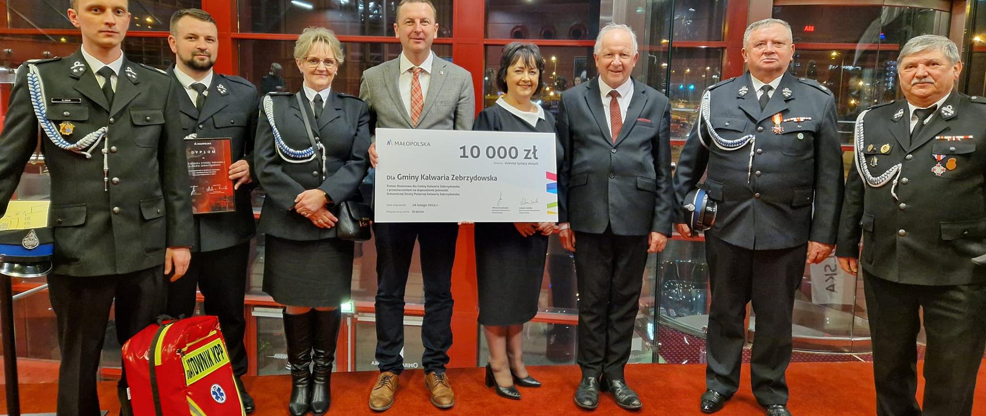 Grupa osób stoi na czerwony dywanie. Pięć osób w strażackich mundurach. Centralnie kobieta i mężczyzna trzymają czek o wartości 10000 zł.