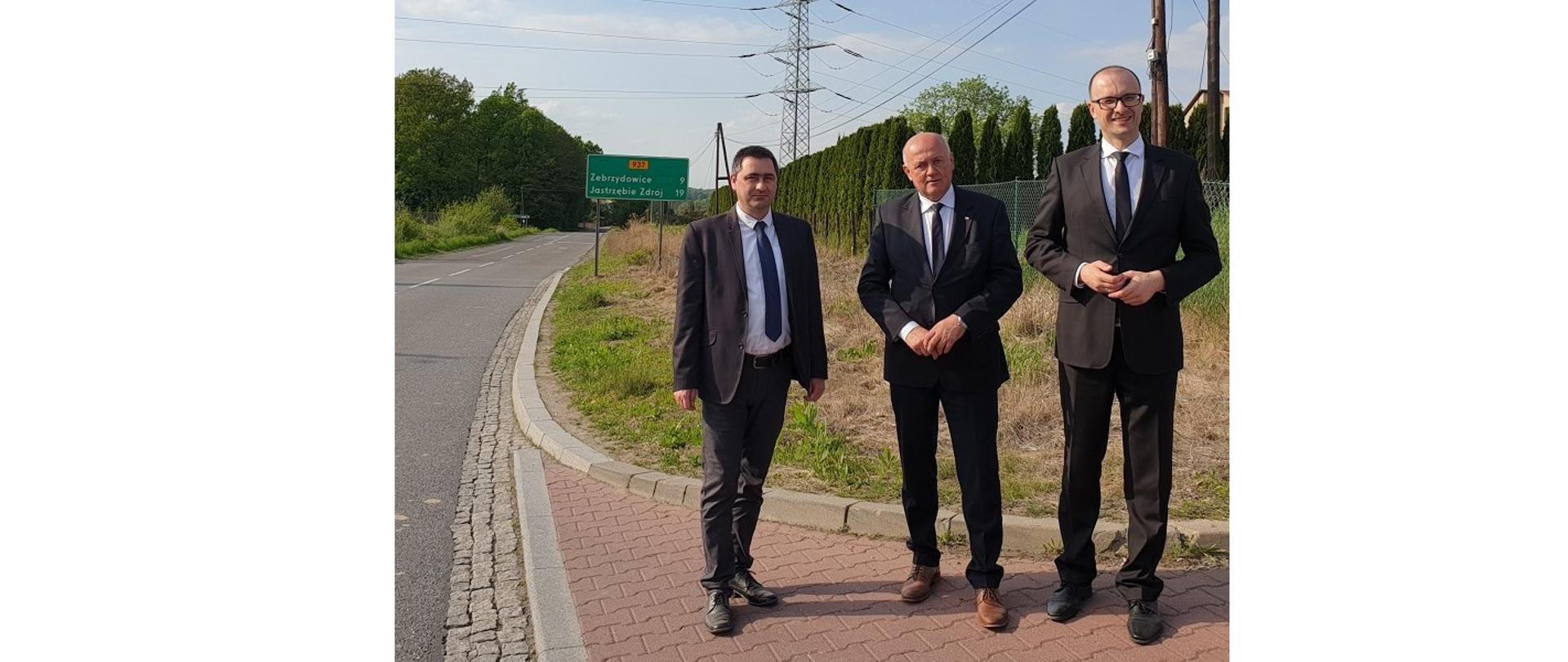 Przy tym odcinku ruchliwej ulicy Jastrzębskiej powstanie kolejny chodnik dla pieszych. Stoją (od lewej): Przewodniczący Rady Gminy Hażlach Sławomir Kolondra, Przewodniczący Sejmiku Województwa Śląskiego Jan Kawulok i Wójt Gminy Hażlach Grzegorz Sikorski. Trzej mężczyźni pozują do zdjęcia. Za nimi z tyłu fragment drogi i przydrożna tablica z podanymi odległościami do najbliższych miejscowości.