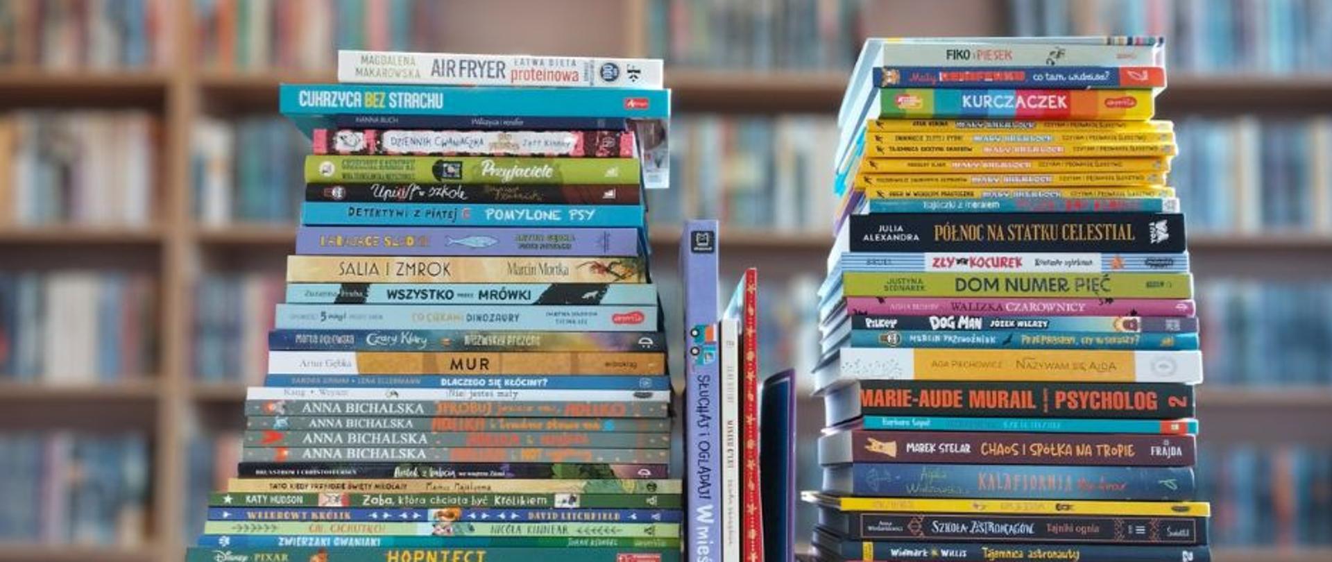 Stos książek dla dzieci i młodzieży ułożonych w dwie wysokie kolumny na stole, w tle rozmyte regały biblioteczne pełne książek.