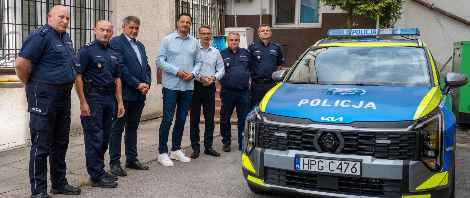 Grupa policjantów i przedstawicieli samorządów stojąca przed budynkiem Komisariatu Policji w Kalwarii Zebrzydowskiej obok nowego, oznakowanego radiowozu marki Kia Sportage.