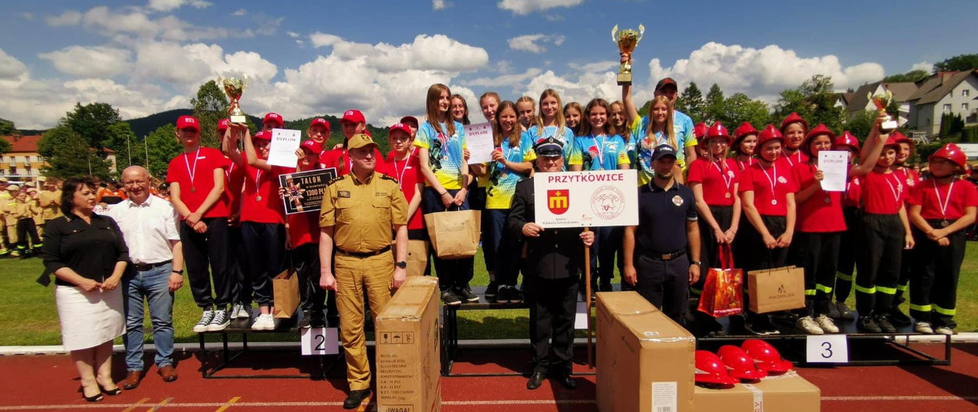 Zwycięskie drużyny młodzieżowe pozują do fotografii grupowej stojąc na podium, przed nimi na bieżni paczki z nagrodami