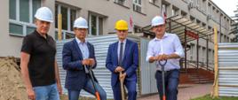 Na zdjęciu przedstawiciele władz pozujący do pamiątkowego zdjęcia na tle prac budowlanych. Panowie mają na głowach ochronne kaski. Trzymają wbite w ziemię łopaty.