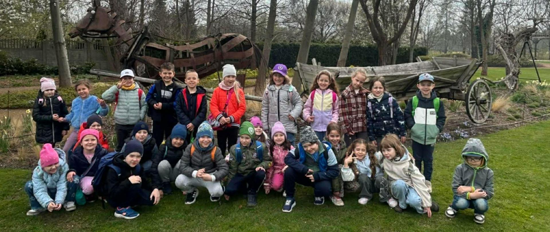 Grupa dzieci pozująca do wspólnego zdjęcia na trawie, w tle rzeźba konia i drewniany wóz w ogrodowej przestrzeni.