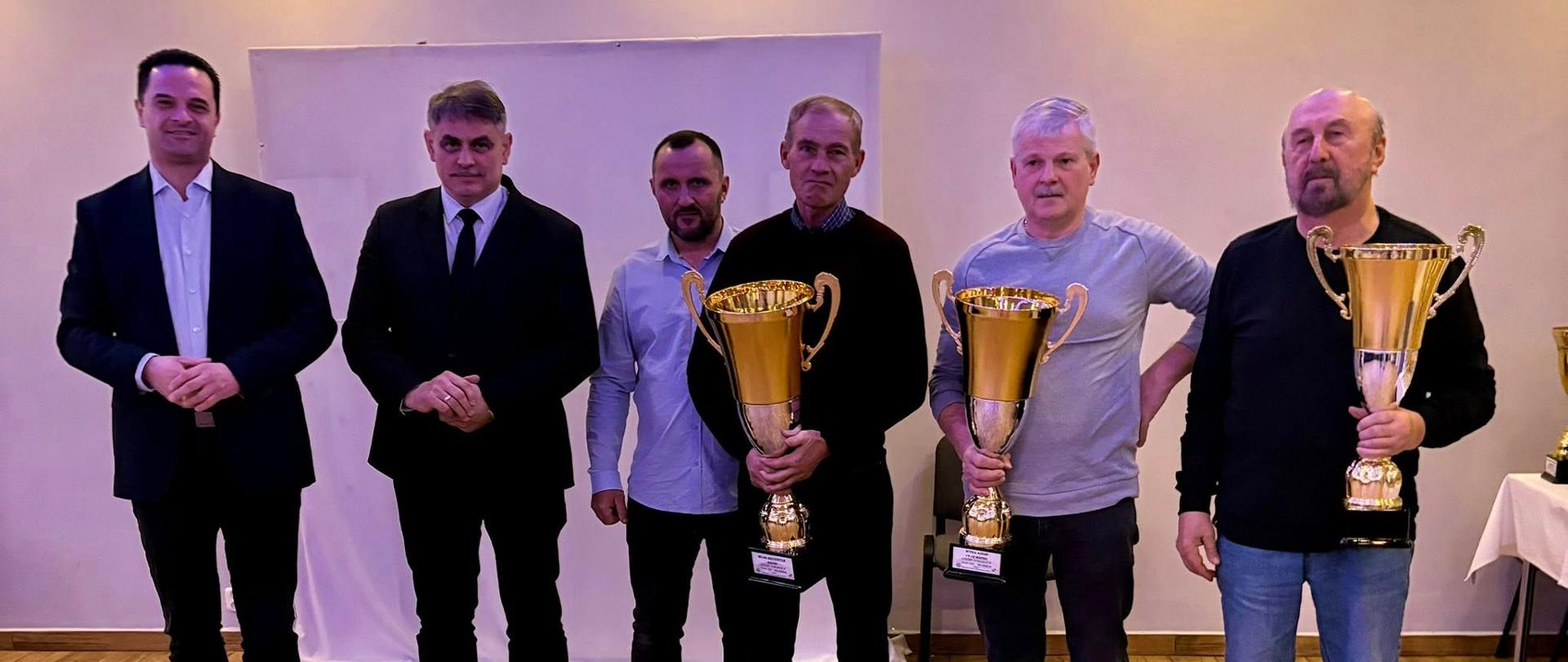 Na zdjęciu widzi się sześciu mężczyzn stojących obok siebie w sali o jasnym oświetleniu. Trzech z nich trzyma duże złote puchary – stoją w prawej części ujęcia. Pozostali trzej stoją po lewej stronie, ubrani elegancko, bez pucharów. Wszyscy patrzą w stronę obiektywu i pozują do wspólnego zdjęcia. Tło jest neutralne, a podłoga wyłożona drewnopodobnymi panelami.
