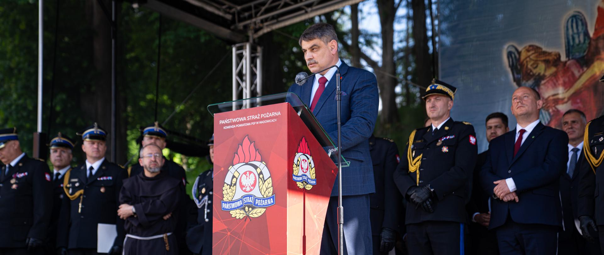Na scenie przy mównicy stoi elegancko ubrany Burmistrz Miasta Kalwarii Zebrzydowskiej,, który przemawia z okazji Obchodów Powiatowego Dnia Strażaka.
W tle widać mężczyzn w strojach strażackich, kustosza Sanktuarium Pasyjno -Maryjnego oraz wojewodę małopolskiego.