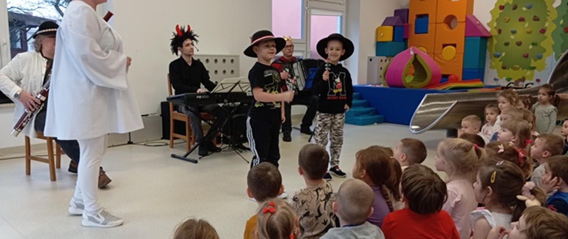 Zdjęcie prezentuje dzieci biorące udział w audycji muzycznej oraz teatrzyku. Przed grupą siedzących dzieci występują aktorzy