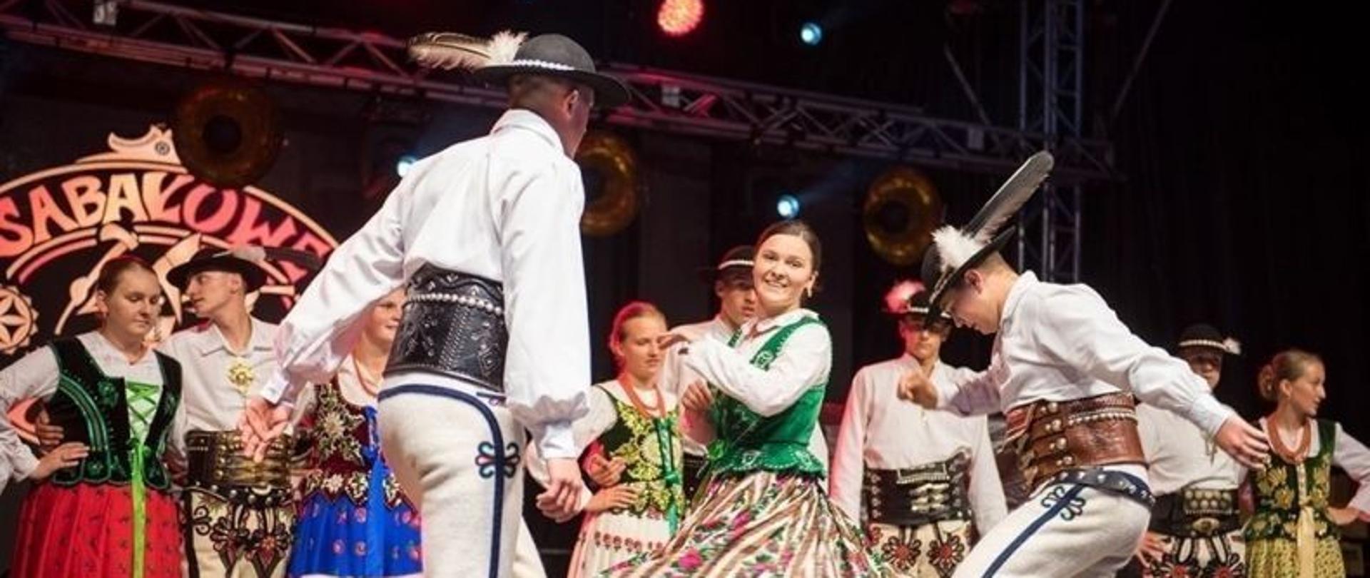 Występ grupy tanecznej, zespołu góralskiego na scenie. Tło występu tworzą pary z zespołu a głównym punktem jest góralka kręcąca się i dwóch górali tańczących koło niej. Wszyscy ubrani w pełne stroje góralskie.