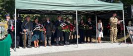 Grupa osób zgromadzona pod zielonym namiotem; widoczne są kwiaty, flagi i elementy uroczystości.