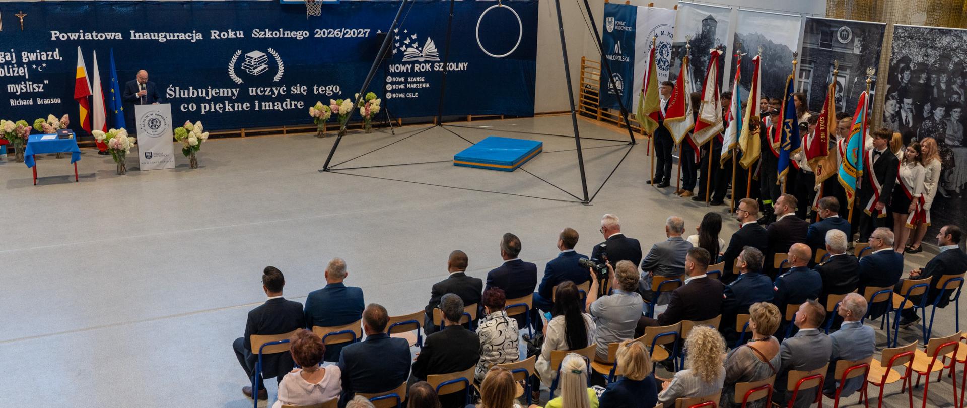 Sala gimnastyczna podczas Powiatowej Inauguracji Roku Szkolnego 2026/2027. Przy mównicy przemawia Burmistrz Miasta Kalwarii Zebrzydowskiej Tadeusz Stela, a zgromadzeni goście oraz poczty sztandarowe uczestniczą w uroczystości.