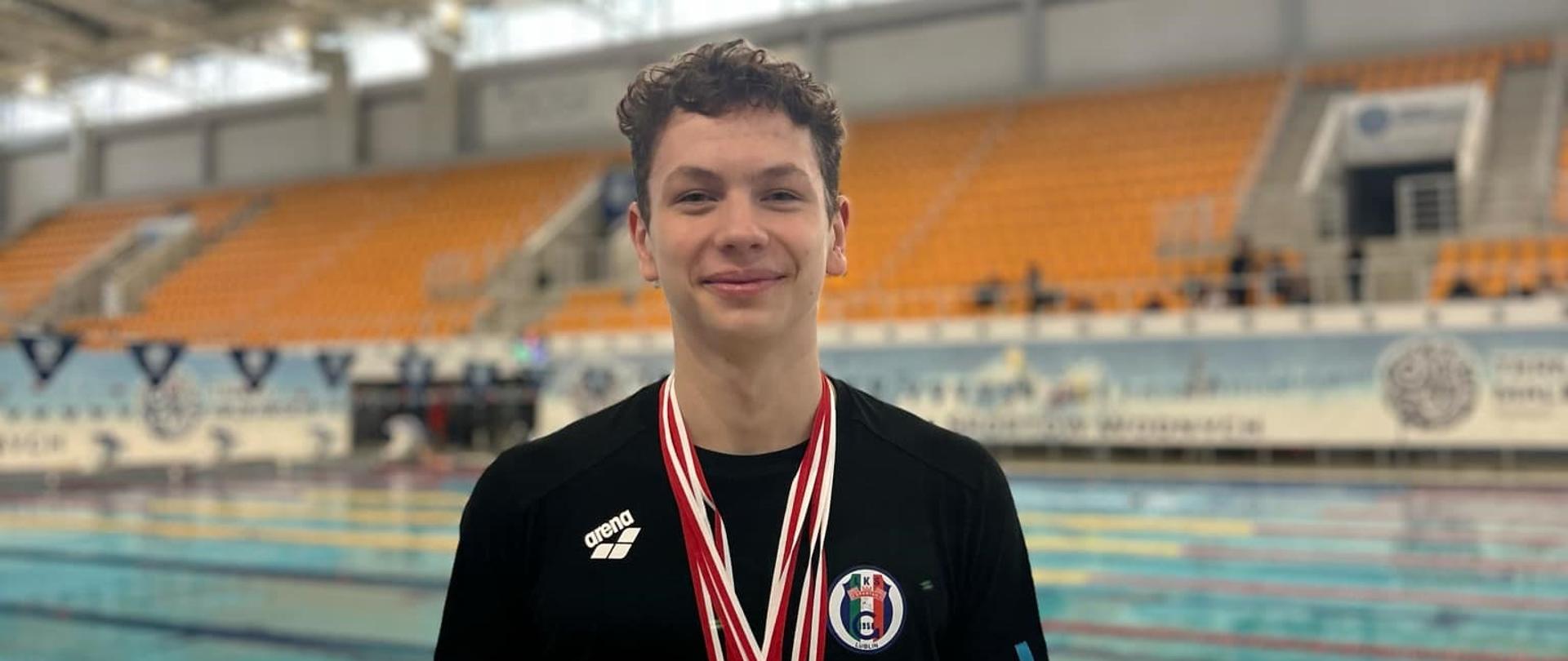 Stojący na tle basenu Rafał Wójcik z zawieszonymi na szyi medalami.