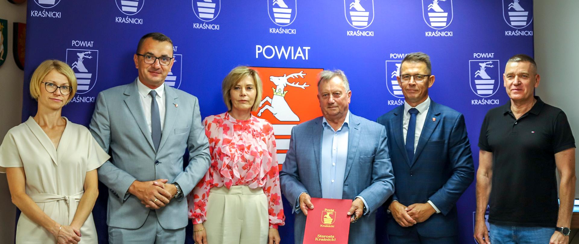 Pamiątkowe zdjęcie z podpisania umowy na realizację inwestycji. Na zdjęciu sześć osób ustawionych w rzędzie obok siebie pozuje na tle ścianki reklamowej powiatu kraśnickiego.
